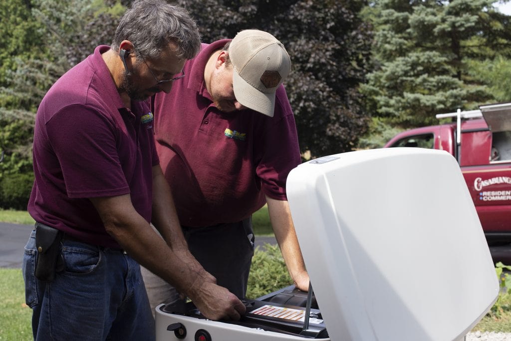 Two male electricians in purple uniforms looking down providing service to a generator